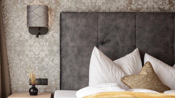 Ein stilvolles Schlafzimmer mit einem grauen Kopfteil und gemusterten Wänden. Auf dem Nachttisch steht eine kleine Lampe und Dekoration. - MYALPS Ötztal - Umhausen
