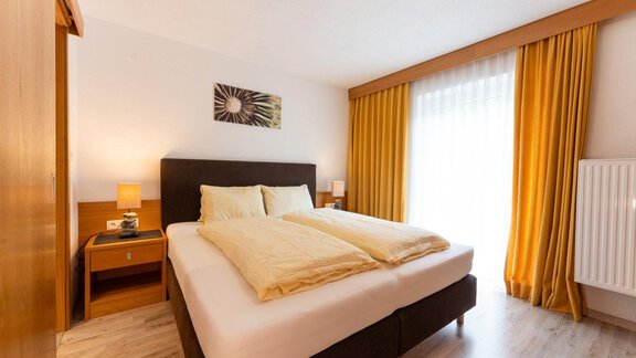 A cozy bedroom with a large bed and bright curtains. The room has a warm wooden tone and plenty of daylight. - Gästehaus Edelweiss - Vent