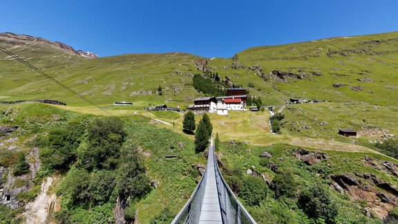 A narrow suspension bridge leads to a building on a green meadow. In the background, gentle hills and a clear blue sky can be seen. - Gästehaus Edelweiss - Vent