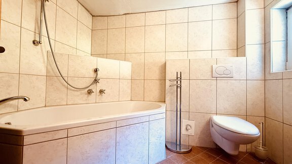 A modern bathroom with a bathtub and a toilet. The walls are tiled and a window provides natural light. - Haus Freistabl - Vent