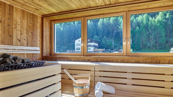 A modern sauna with wooden walls and large windows that provide a beautiful view of the forest. In the foreground, there is a sauna bench, stones, and a wooden container. - Hotel Vent - Vent