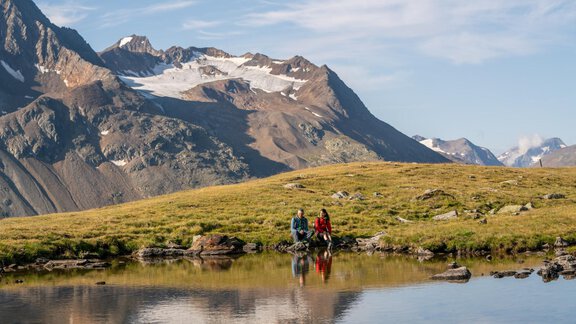 A picturesque mountain landscape with snow-covered peaks and a calm lake. Two people are sitting by the shore and enjoying the view. - Hotel Vent - Vent
