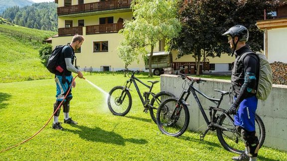 Two mountain bikers are standing on a green meadow next to their bikes. One of the bikers is cleaning his bike with a high-pressure cleaner in front of an accommodation building. - Apart Praxmarer - Zwieselstein
