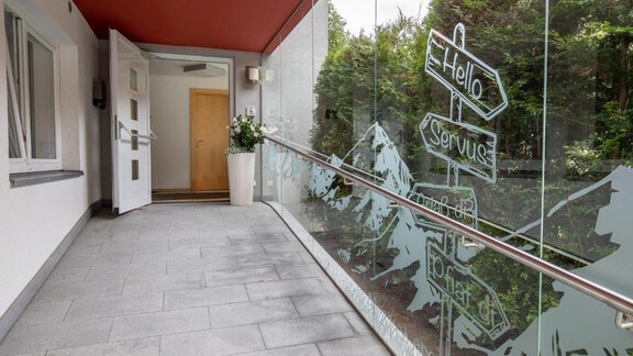 A modern entrance area with glass walls and a plant. On the glass wall, drawings of mountains and signposts can be seen. - Apart Praxmarer - Zwieselstein
