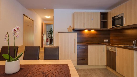 A modern kitchen with wooden cabinets and stainless steel appliances. A table with a flower and chairs is in the foreground. - Apart Praxmarer - Zwieselstein