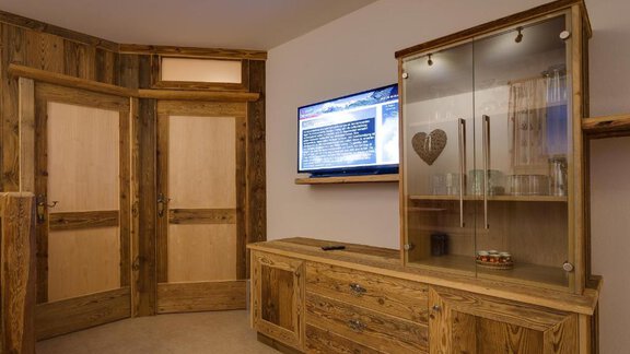 A modern room with wood paneling and a large television on the wall. A beautiful cabinet with glass doors stands next to the two entrance doors. - Apart Praxmarer - Zwieselstein