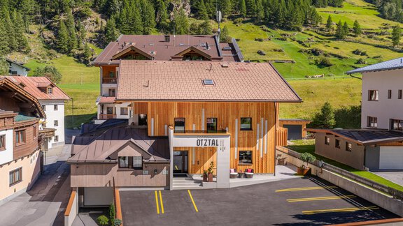 A modern building with a wooden facade amidst green mountains. In the foreground, there is a parking lot with marked parking spaces. - Garni Ötztal ROOMS & APARTS - Zwieselstein