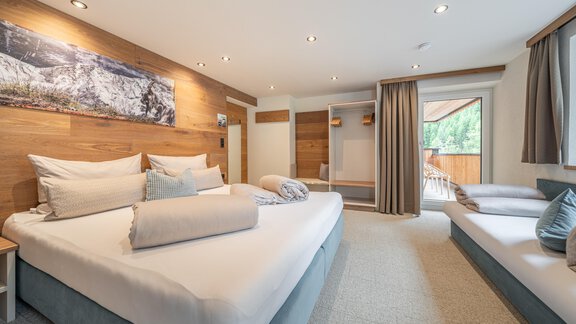 A modern hotel room with a large double bed and an additional single bed. The walls are in a warm wood tone and there is plenty of daylight through the windows. - Garni Ötztal ROOMS & APARTS - Zwieselstein