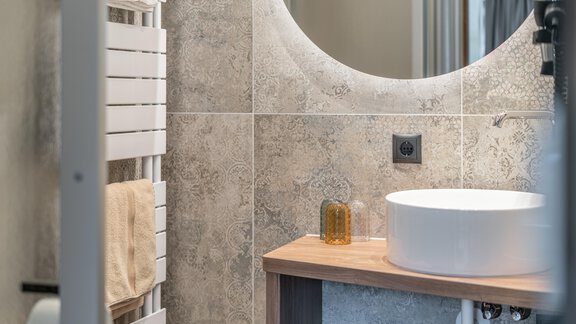 A modern bathroom with elegant tiles and a round mirror. Beneath it is a sink made of white ceramic and a wooden shelf. - Garni Ötztal ROOMS & APARTS - Zwieselstein