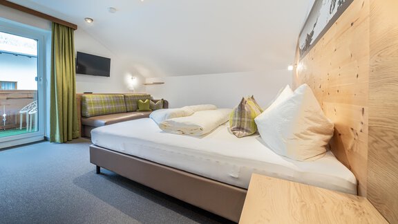 A modern bedroom with a large bed and cozy pillows. There is a small balcony and a seating area with a television. - Garni Ötztal ROOMS & APARTS - Zwieselstein