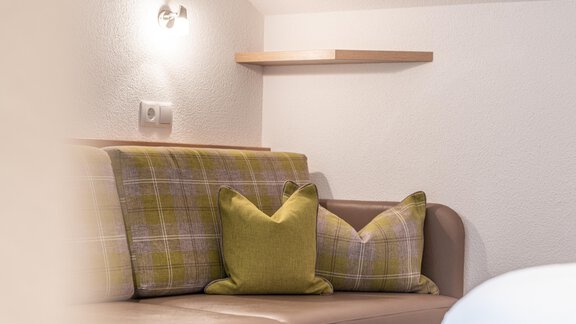 A cozy seating area with a brown sofa and green cushions. Next to the sofa, there is a shelf and wall lighting. - Garni Ötztal ROOMS & APARTS - Zwieselstein