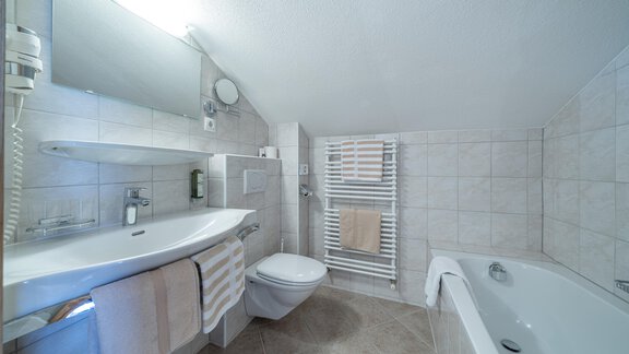 A modern bathroom with a bathtub and a shower. The walls are outfitted with light tiles and there is a large mirror above the sink. - Garni Ötztal ROOMS & APARTS - Zwieselstein