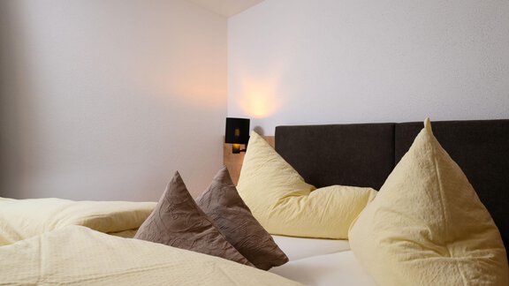 A cozy bedroom with a dark bed frame and soft pillows. The walls are light, and a warm glowing lamp provides an inviting atmosphere. - Gästehaus Judith - Zwieselstein