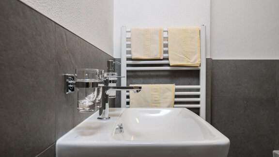 A modern bathroom with a small sink and towels on a radiator. The walls are tiled in light and dark colors. - Gästehaus Judith - Zwieselstein
