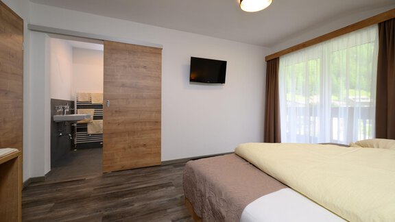 A modern room with two beds and a flat-screen TV. In the background, a well-equipped bathroom is visible. - Gästehaus Judith - Zwieselstein