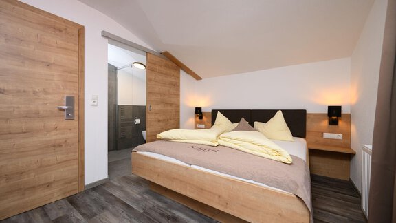 A modern bedroom with a double bed, bright pillows, and wooden accents. The walls are plain, and the room feels inviting and cozy. - Gästehaus Judith - Zwieselstein
