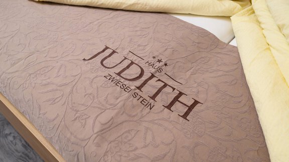 A bed with decorative bedding in beige and a yellow blanket. The bedding features the inscription "Haus Judith" in an elegant font. - Gästehaus Judith - Zwieselstein