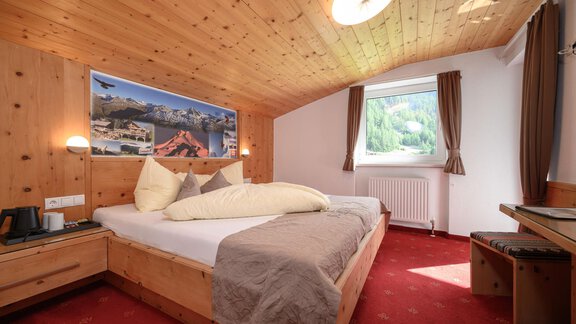 A cozy bedroom with wooden walls and a large window. The bed is equipped with pillows and a blanket, and a beautiful view of nature is visible. - Gästehaus Judith - Zwieselstein