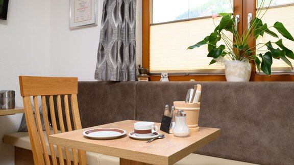 A cozy dining area with a wooden table and a wooden bench. On the table are dishes and cutlery, as well as a plant in the background. - Gästehaus Judith - Zwieselstein