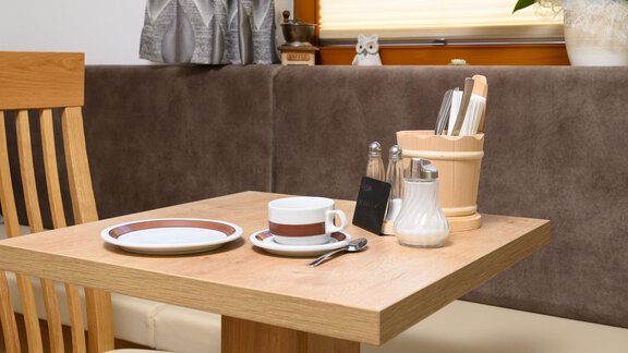 A table with a coffee mug, a plate, and cutlery. In the background are utensil holders and a plant. - Gästehaus Judith - Zwieselstein