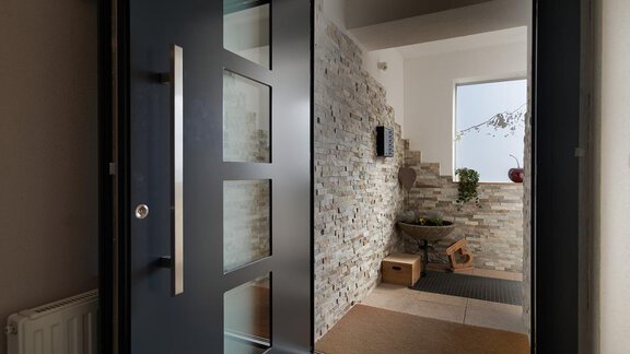 A modern entrance area with a stylish door. On the left side, there are walls with natural stone and a bright window front. - Gästehaus Judith - Zwieselstein