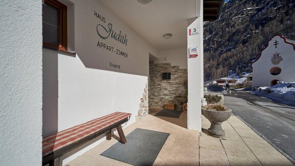 An inviting entrance area with a bench and a modern façade. In the background, a picturesque mountain landscape and a small chapel can be seen. - Gästehaus Judith - Zwieselstein