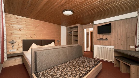 A cozy room with wooden paneling, a double bed, and a seating area. There is a television on the wall and a desk in the corner. - Gästehaus Judith - Zwieselstein