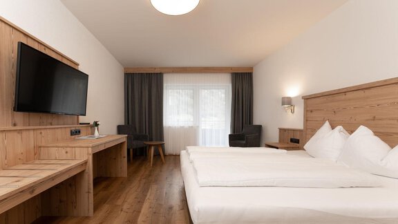 A modern hotel room with a double bed and wood paneling. There is a television and a desk as well as a window with curtains. - Gasthof Brückenwirt - Zwieselstein