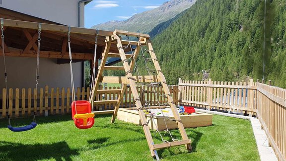 A beautiful playground with a wooden swing and a slide. In the background, green mountains and a blue sky can be seen. - Haus Fortuna - Zwieselstein