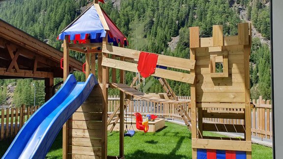 A playground with a slide and a small wooden tower. The area is surrounded by a wooden border and has a lot of green grass. - Haus Fortuna - Zwieselstein