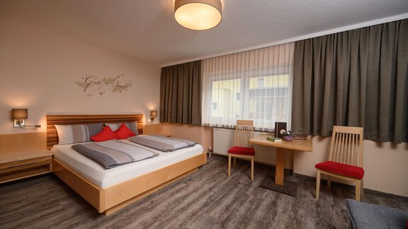 A modern hotel room with a large bed, wooden furniture, and a cozy workspace. The windows are equipped with curtains and the room is well lit. - Haus Sonnschein - Zwieselstein
