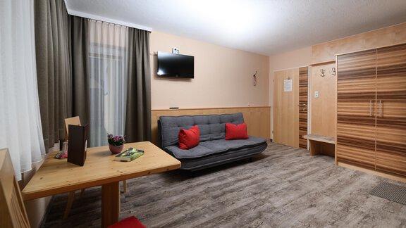 A cozy living space with a sofa bed and a wooden table. The walls are light, and the room has large windows with curtains. - Haus Sonnschein - Zwieselstein