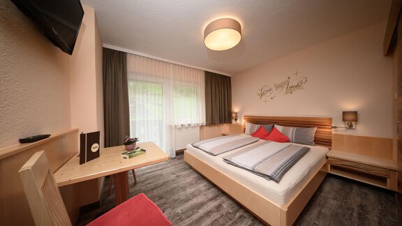 A modern room with a double bed, bright walls, and wooden details. There is a table with a chair and windows that let in plenty of daylight. - Haus Sonnschein - Zwieselstein