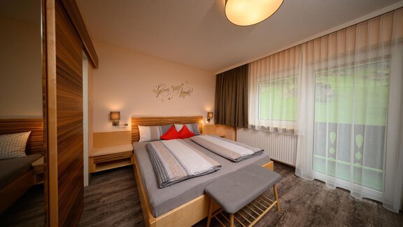 A modern bedroom with a large bed and light curtains. Next to the bed is a small stool and there is a wall shelf. - Haus Sonnschein - Zwieselstein
