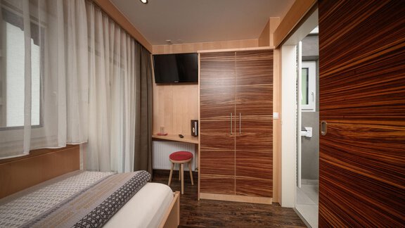 A modern hotel room with a single bed and a desk. The walls are designed in warm wood tones, and the room has large windows with curtains. - Haus Sonnschein - Zwieselstein