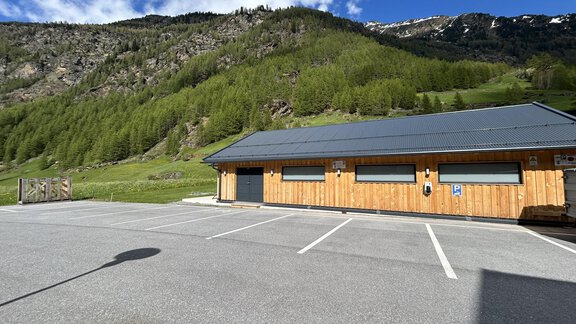 A wooden building with a modern roof stands in a quiet environment. In the background, green mountains and a blue sky can be seen. - Hochstatt Appartements - Zwieselstein