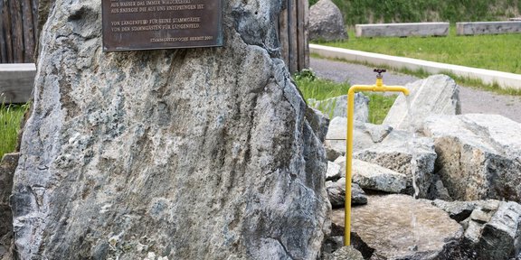 A large rock with a metal plaque and a yellow faucet is located in a green environment. The rock is surrounded by stones and creates a clear focal point. - Thermal-Heilwasser Badl' Längenfeld - Längenfeld