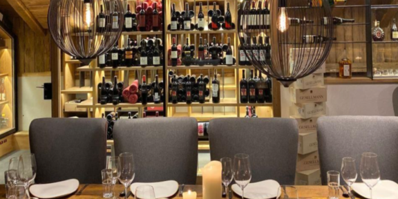 Ein elegantes Restaurant mit einem großen Holztisch und stilvollen Stühlen. Im Hintergrund befindet sich eine Wand mit einer Auswahl an Weinen und modernen Hängeleuchten. - Hühnersteign - Sölden