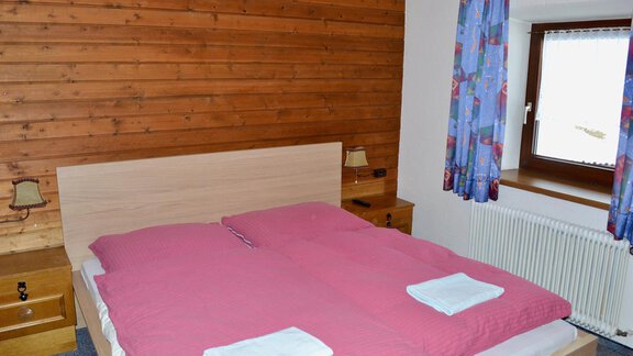 A cozy bedroom with a large bed and pink bedding. The walls are made of wood, and there are windows with colorful curtains. - Haus Maria - Hochsölden