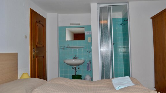 A simple room with a bed, a sink, and a shower. The walls are light, and there is a wooden wardrobe. - Haus Maria - Hochsölden