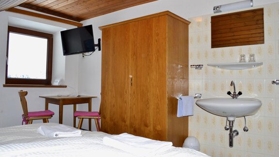 A simple room with a double bed, a wooden wardrobe, and a TV on the wall. There is a table with two chairs and a sink in the corner. - Haus Maria - Hochsölden