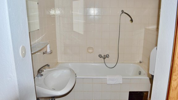 A simple bathroom with a bathtub, a sink, and a shower. The walls are light-tiled and the furnishings are functional. - Haus Maria - Hochsölden