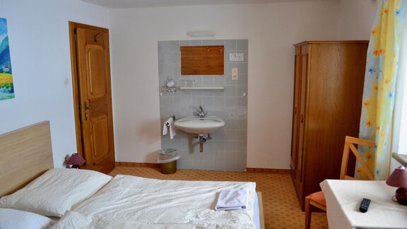 A simple room with a double bed, a table, and a chair. The walls are light, and there is a small washing unit in the corner. - Haus Maria - Hochsölden