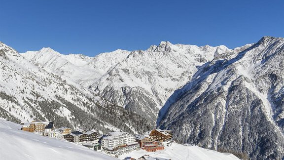 A picturesque mountain landscape with snow-covered peaks and a clear blue sky. In the foreground, several houses can be seen in the winter scenery. - Pension Ferdinand - Hochsölden