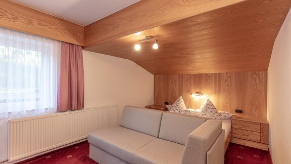 Ein gemütliches Zimmer mit einem grauen Sofa und einem Doppelbett. Die Wände sind aus Holz und es gibt ein großes Fenster, das für Tageslicht sorgt. - Apart Felsenhof - Huben