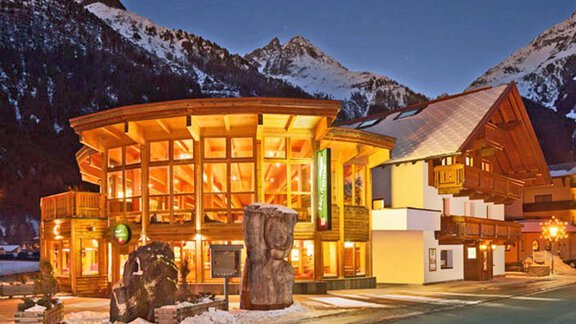 A modern wooden building in the Alps at night. Surrounded by snow-covered mountains, it radiates an inviting atmosphere. - Daheim Appartements - Huben