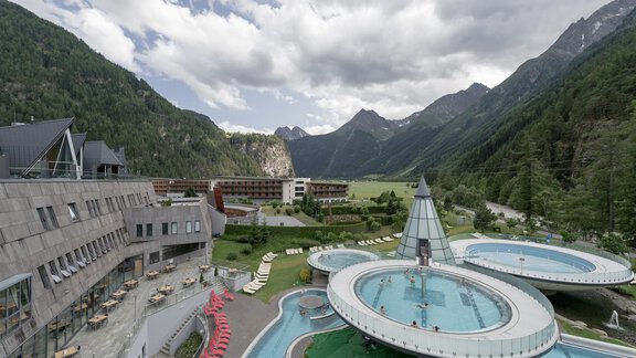 A modern hotel with a wellness area and several swimming pools, surrounded by impressive mountains. The sky is partly cloudy and the landscape appears inviting and tranquil. - Daheim Appartements - Huben