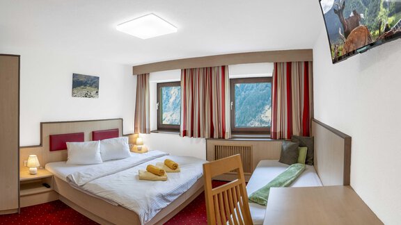 A cozy hotel room with two beds and a single bed. The windows are decorated with striped curtains and there is a small table with a plant. - Berghof Reich - Längenfeld