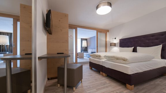 A modern bedroom with a large bed and stylish decor. The room is bright and cozy, with a table and a television. - Hotel  Bergwelt - Längenfeld