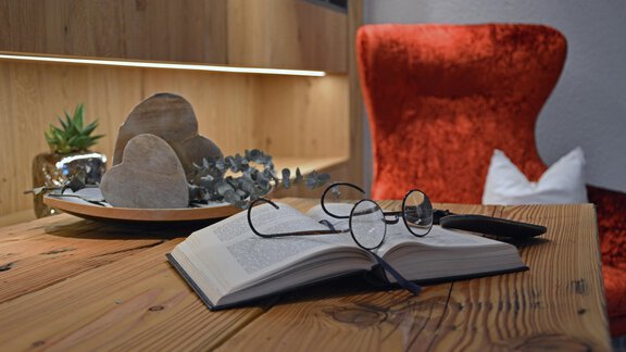 Ein aufgeschlagenes Buch liegt auf einem Holztisch. Neben dem Buch befinden sich eine Lesebrille und einige dekorative Gegenstände auf einem Teller. - Hotel Stern - Längenfeld